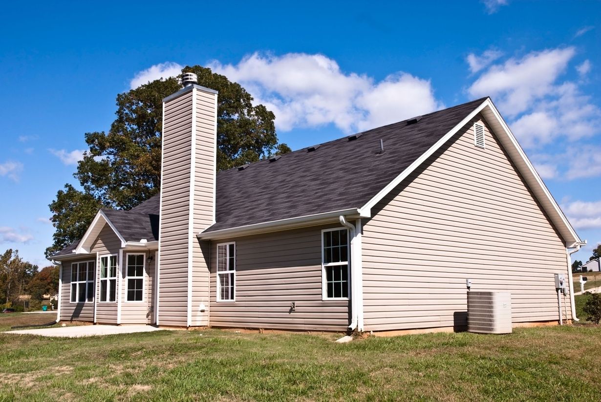 A single-story beige house with a tall stone chimney, dark shingled roof, and green lawn under a bright blue sky.