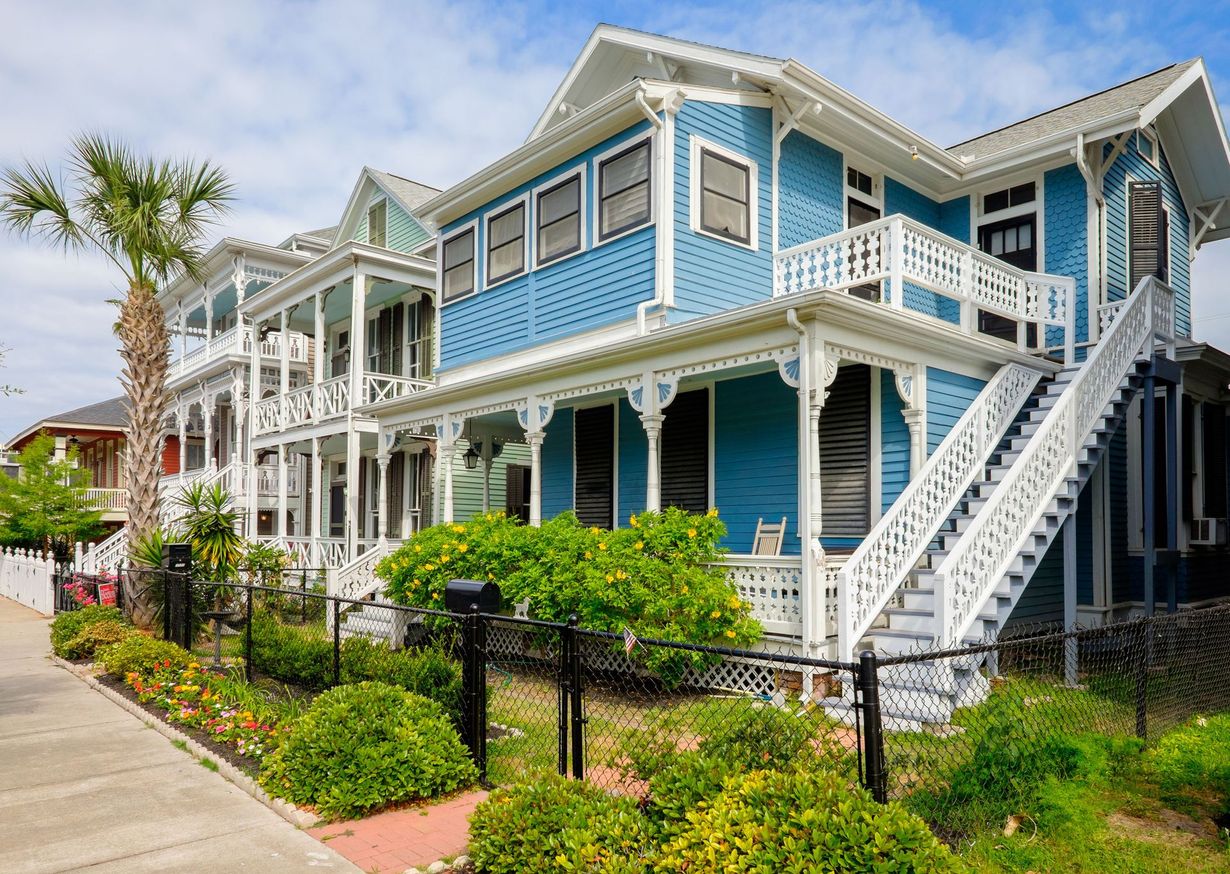 A row of blue, two-story Victorian-style houses with white porches and stairs, seen from a sidewalk on a sunny day.