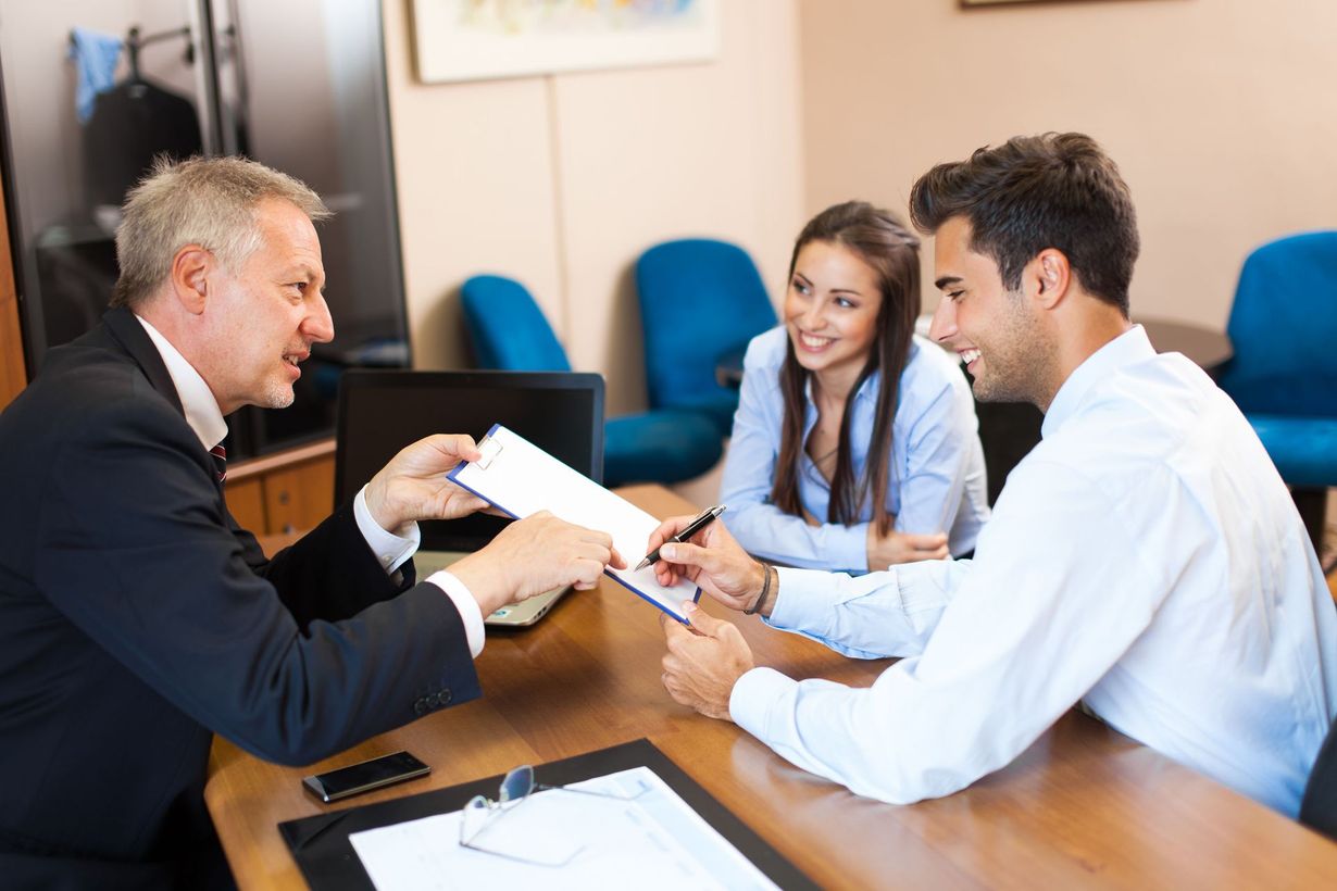 An advisor sits across from a couple at a wooden desk, gesturing toward a contract for them to sign in an office.