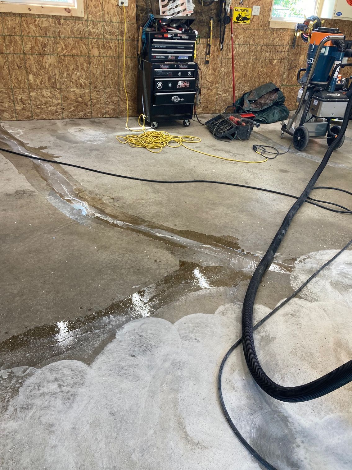 Garage floor being cleaned with a hose, showing suds and wet patches. A tool chest and power tool are visible.
