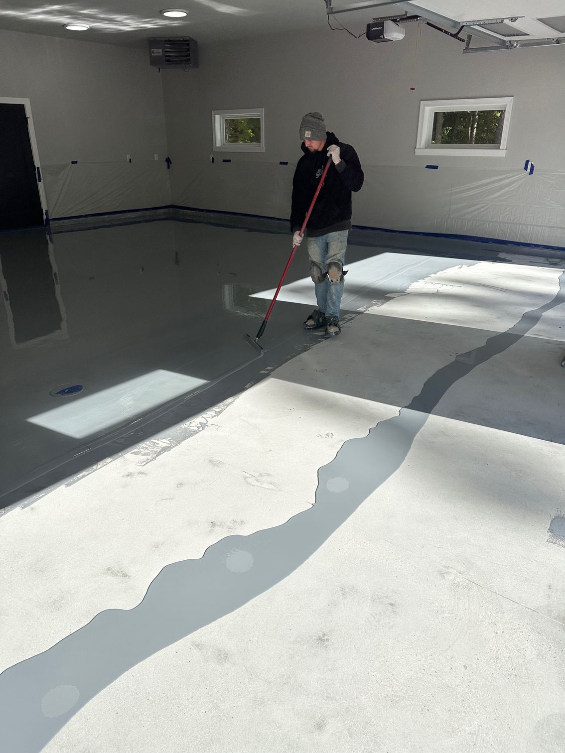 Person applying epoxy coating to a garage floor with a long-handled squeegee. Gray floor, blue tape, and white walls.