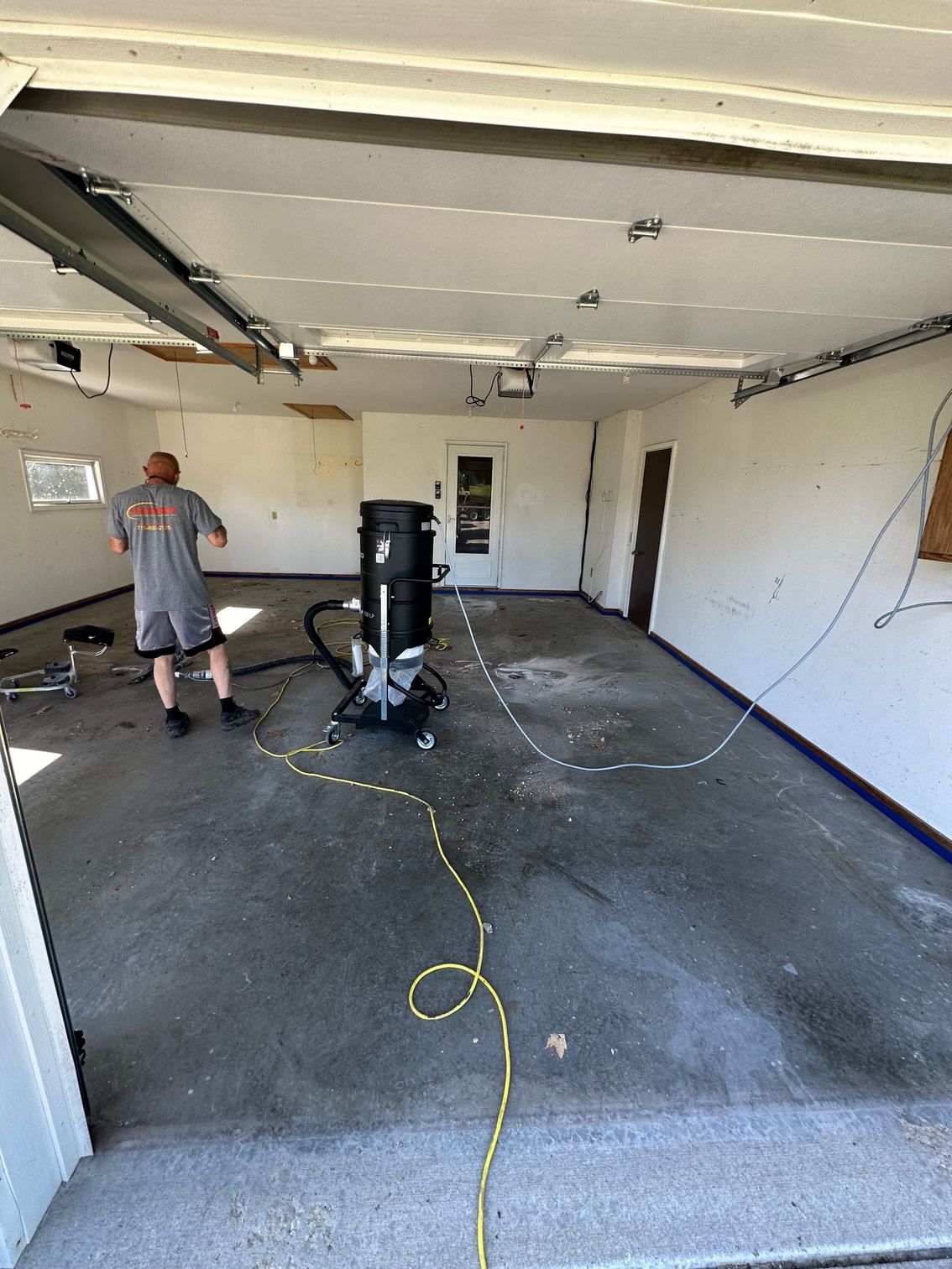 Person vacuuming a garage floor with a large industrial vacuum. Garage has exposed ceiling beams and an open doorway.