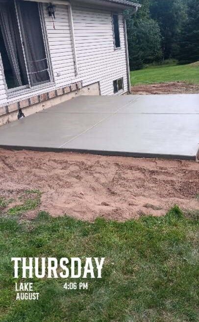 Newly poured concrete patio next to a house with a green lawn and trees in the background.