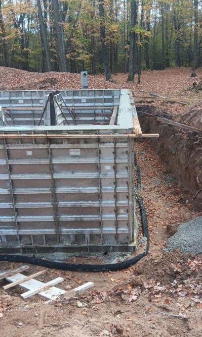 Construction site with concrete forms in place for a foundation. Soil, trees, and gravel visible.