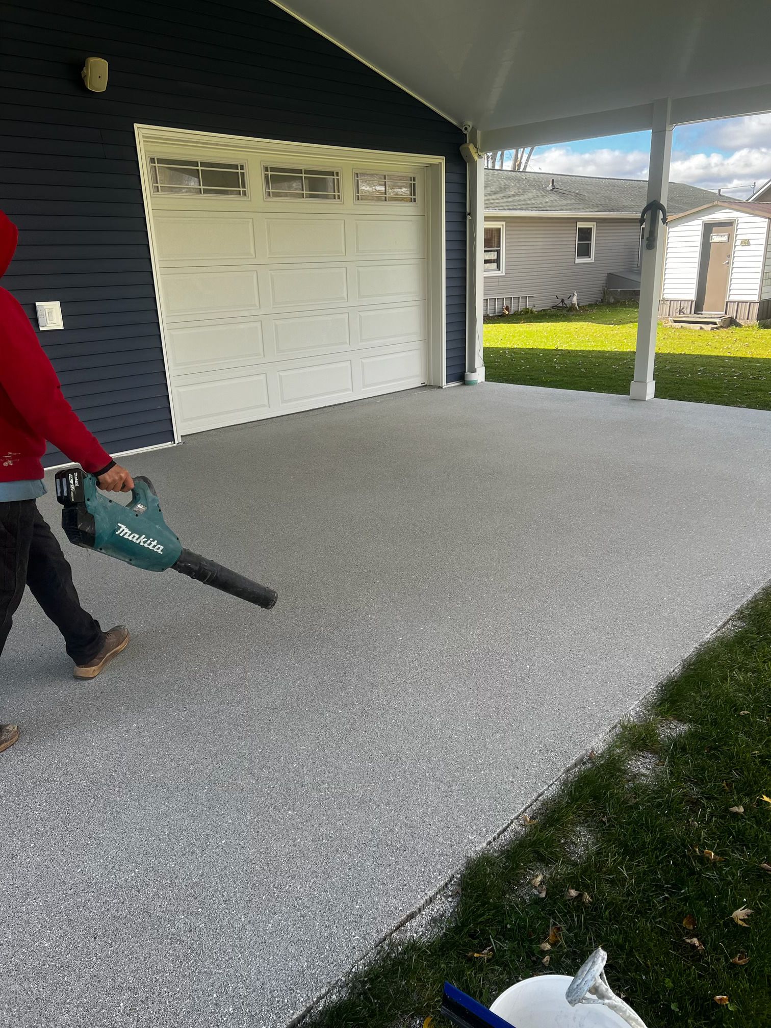 Person using a leaf blower on a gray concrete patio, next to a garage and lawn.