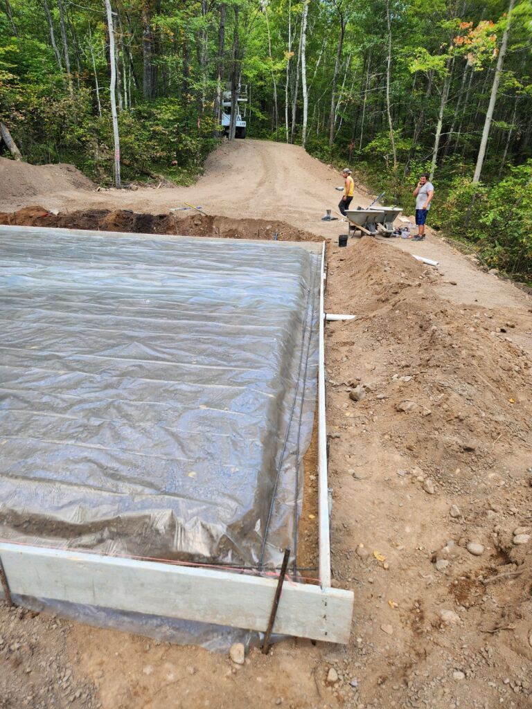 Construction site with concrete foundation covered in plastic. Two workers nearby; dirt road leads to woods.