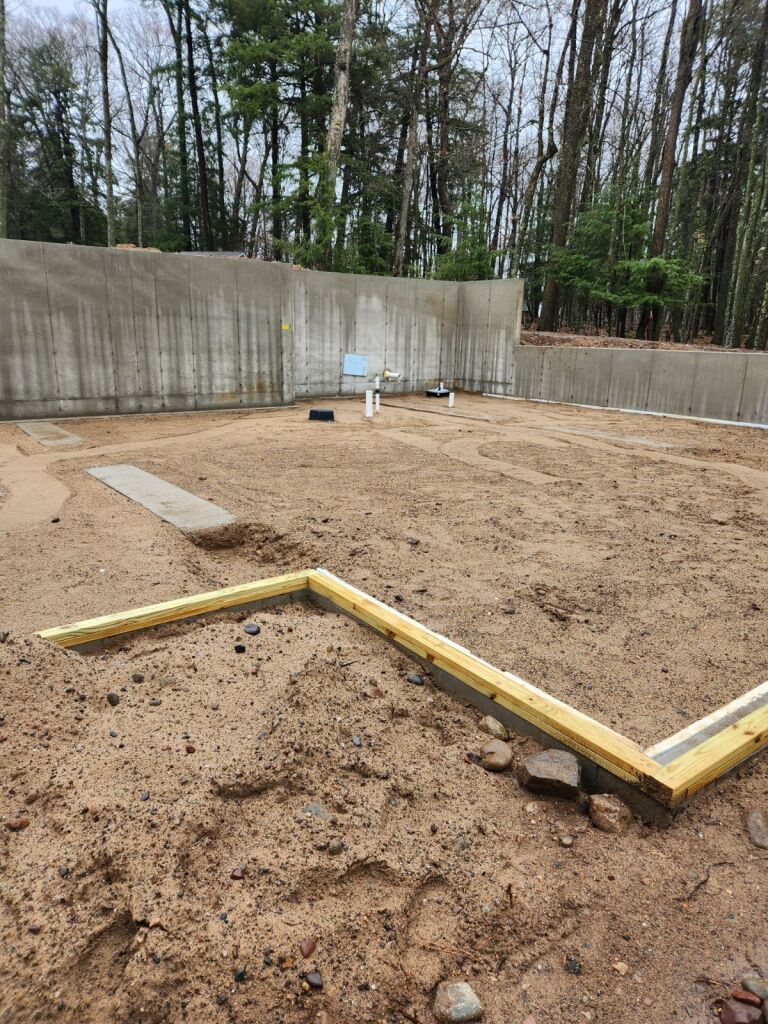 Construction site with concrete foundation walls and sand-covered ground; a yellow framed structure outlines a future wall.