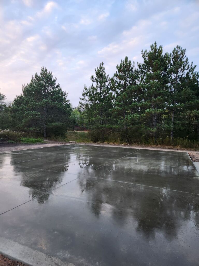 Wet concrete area reflecting trees under a cloudy sky.