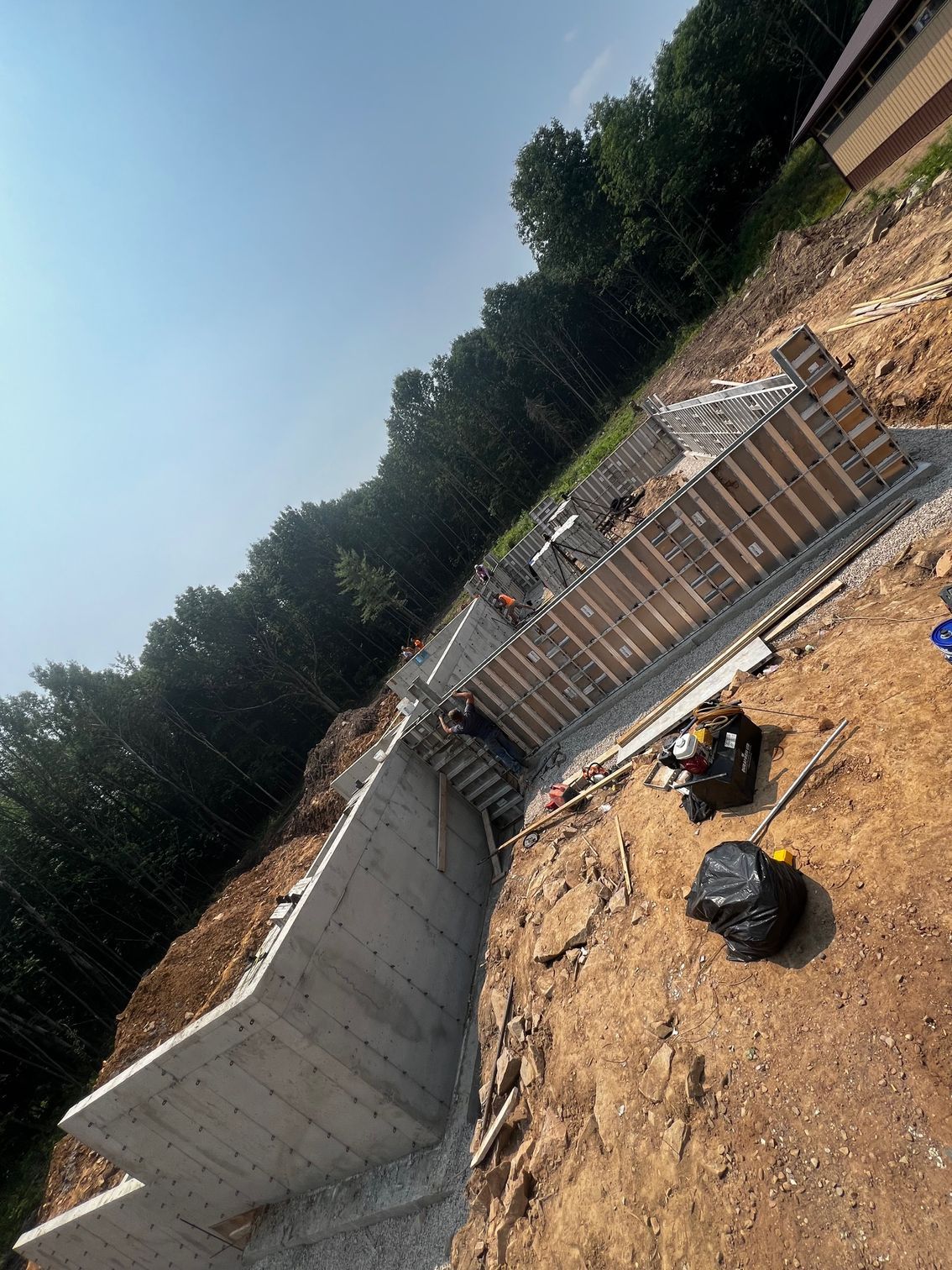 Construction site with concrete retaining walls on a hillside. Dirt and trees surround the structure.