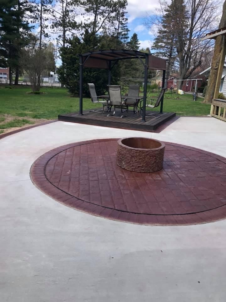 Concrete patio with a circular brick-patterned area around a fire pit and a pergola in the background.