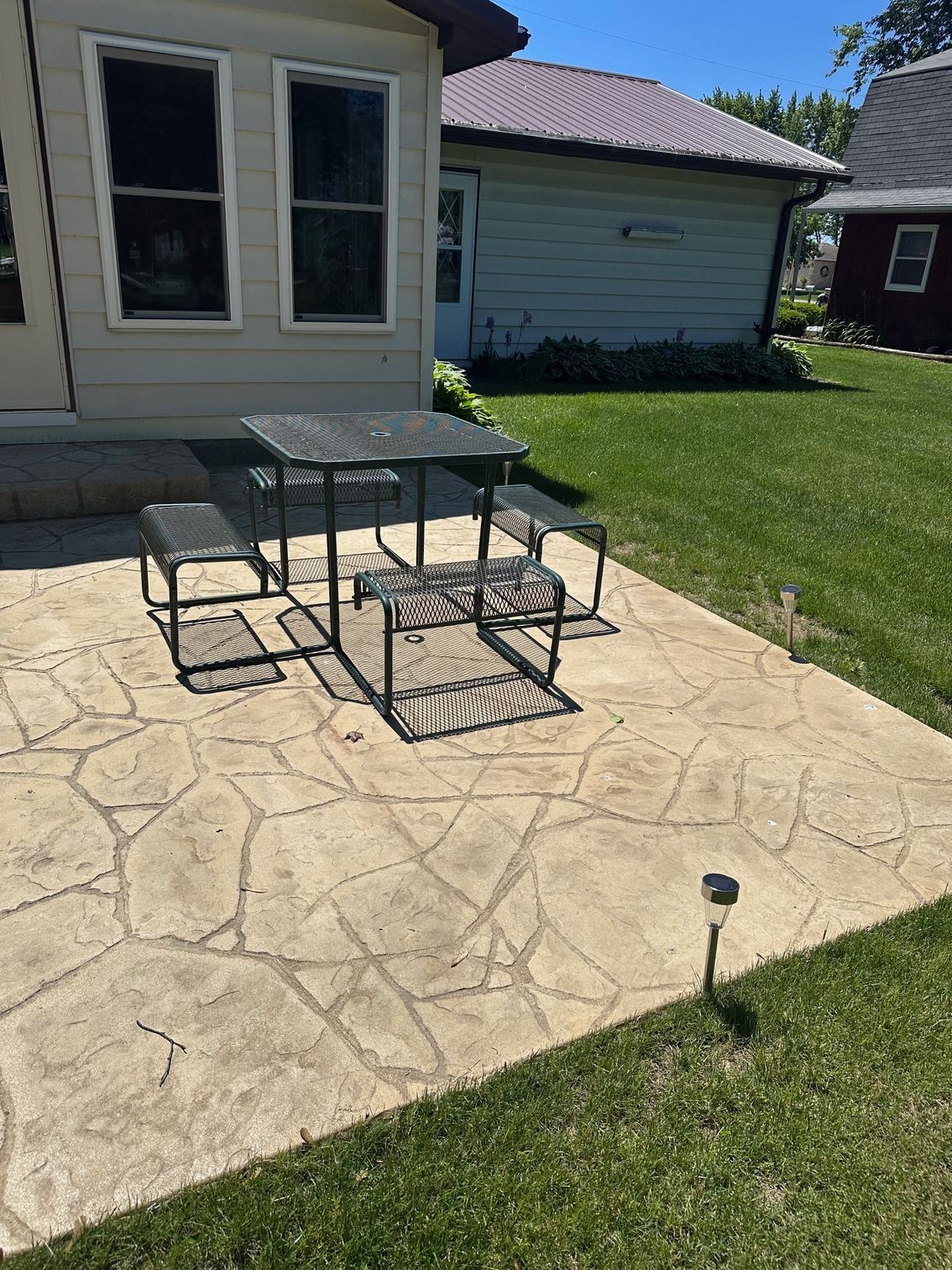 Patio with metal furniture on stamped concrete, beside a building and grassy lawn.