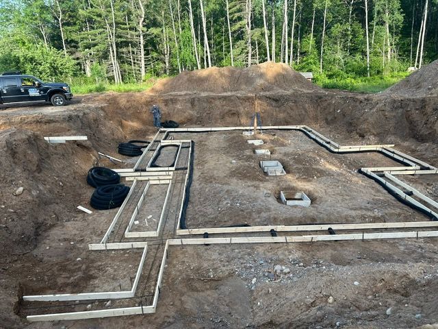 Foundation excavation for a building with wooden forms and pipes visible, outdoors.