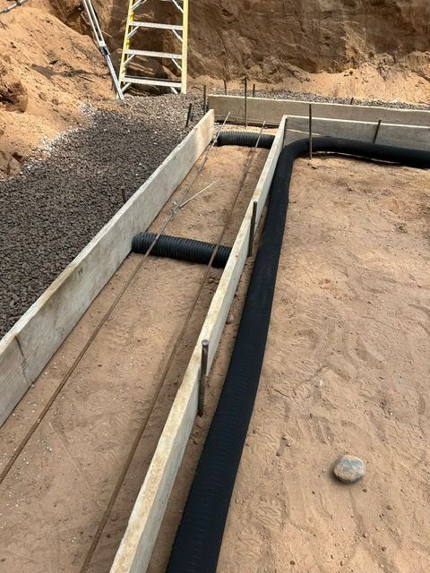 Construction site: concrete foundation forms with rebar, drain pipe, and gravel on dirt ground.