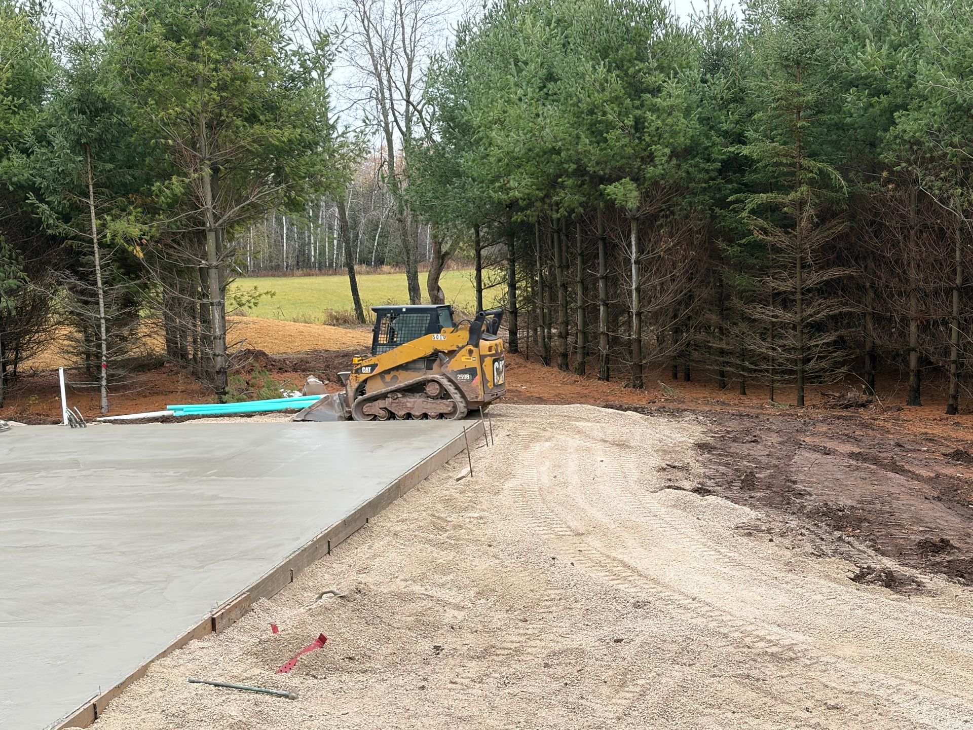 A small yellow skid steer compacting soil next to a newly poured concrete pad, with trees in the background.