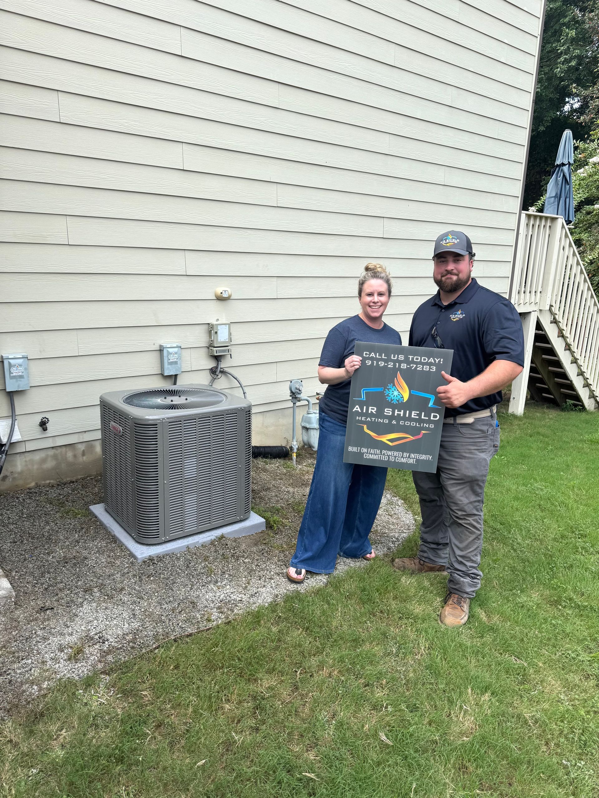 Two people pose with a sign next to a new AC unit outside a house.