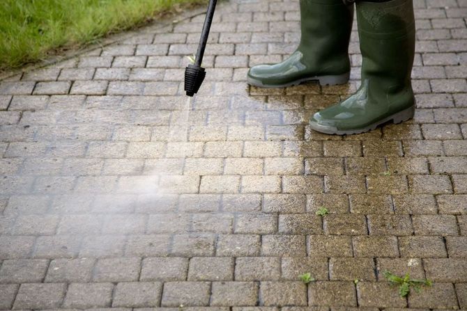 Person wearing green boots sprays a white substance onto a brick sidewalk.