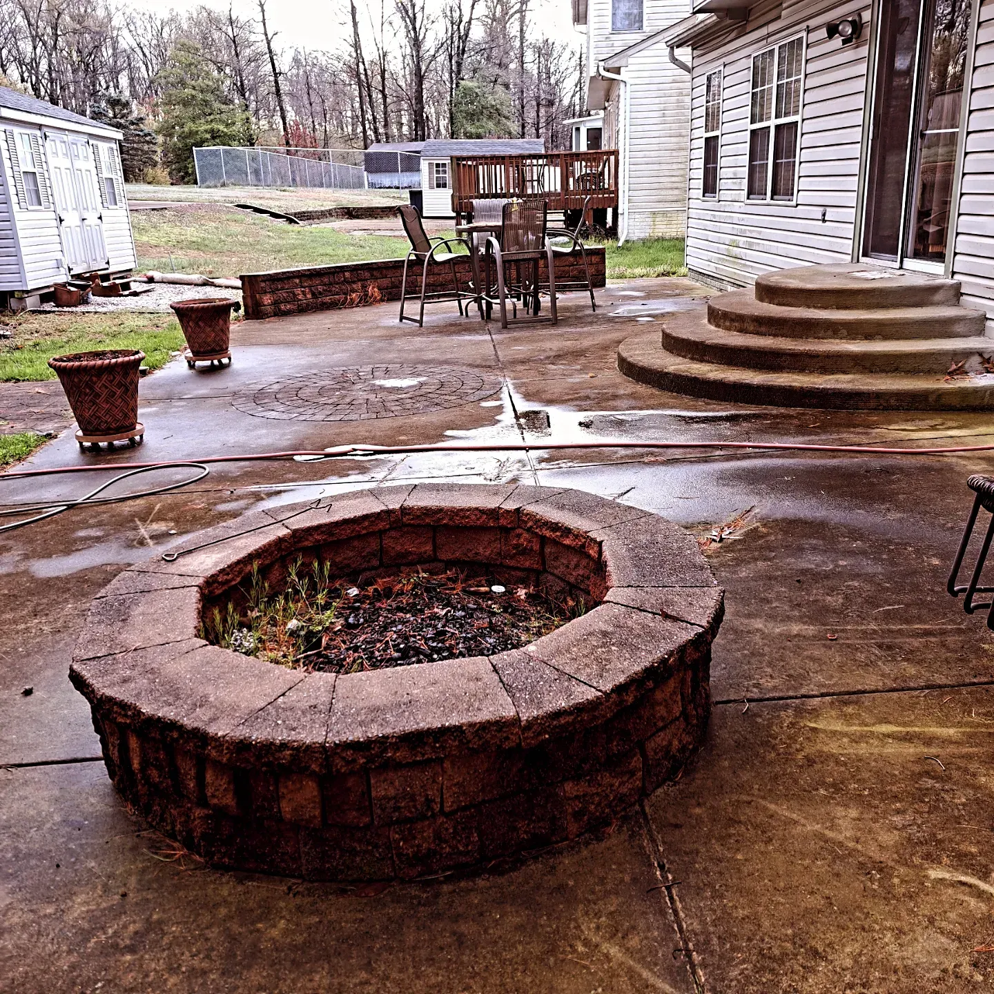 Backyard patio with a brick fire pit, wet concrete, potted plants, and steps leading to a house.