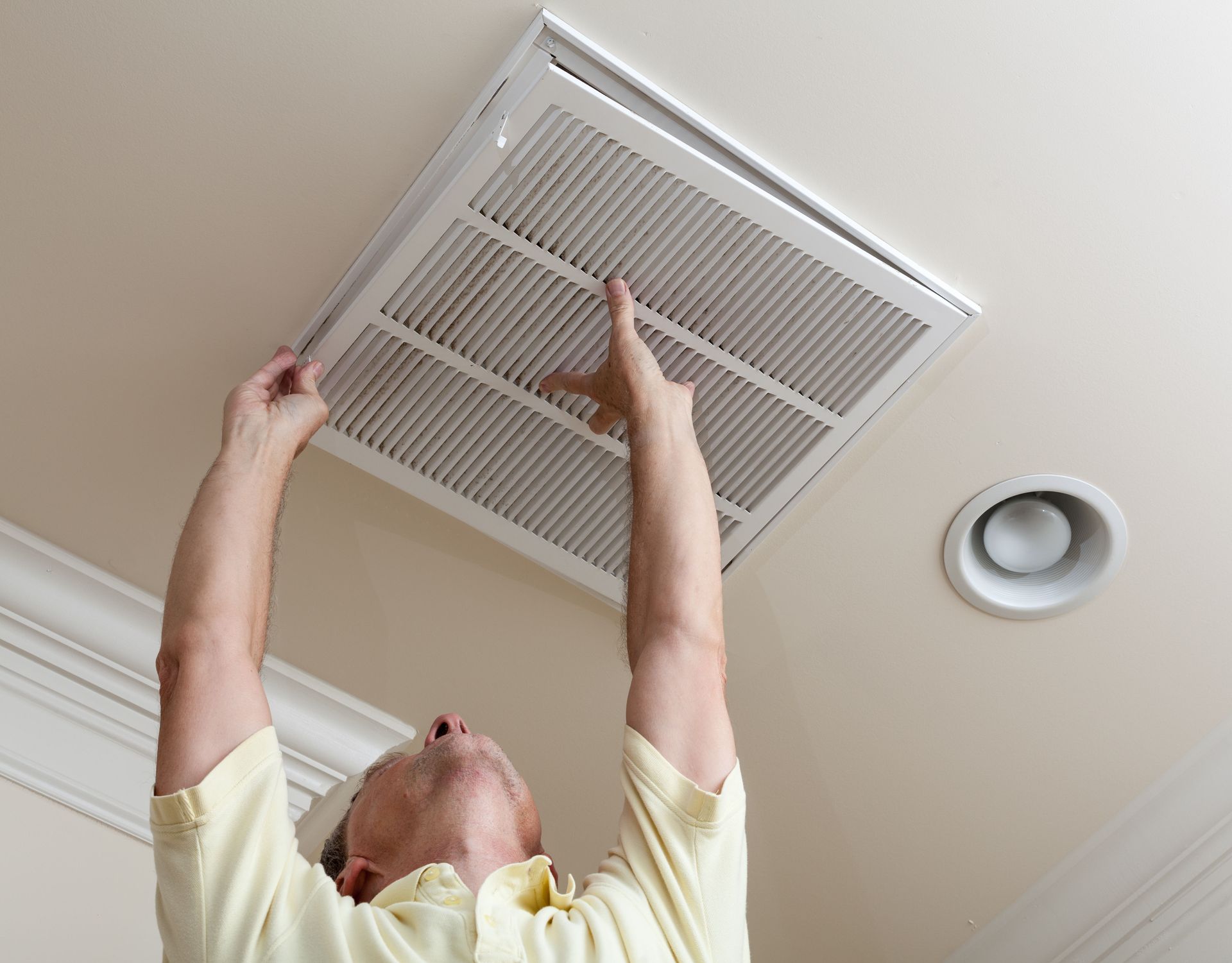 A person in a light yellow shirt standing on a ladder, reaching up to replace a square HVAC air filter in a ceiling vent.