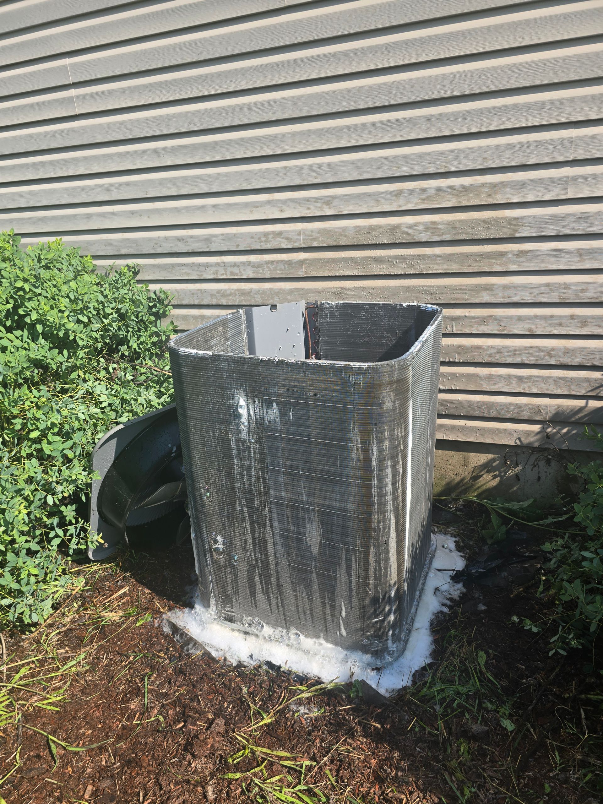 Outdoor HVAC unit wrapped in silver insulation beside a house wall and shrubs
