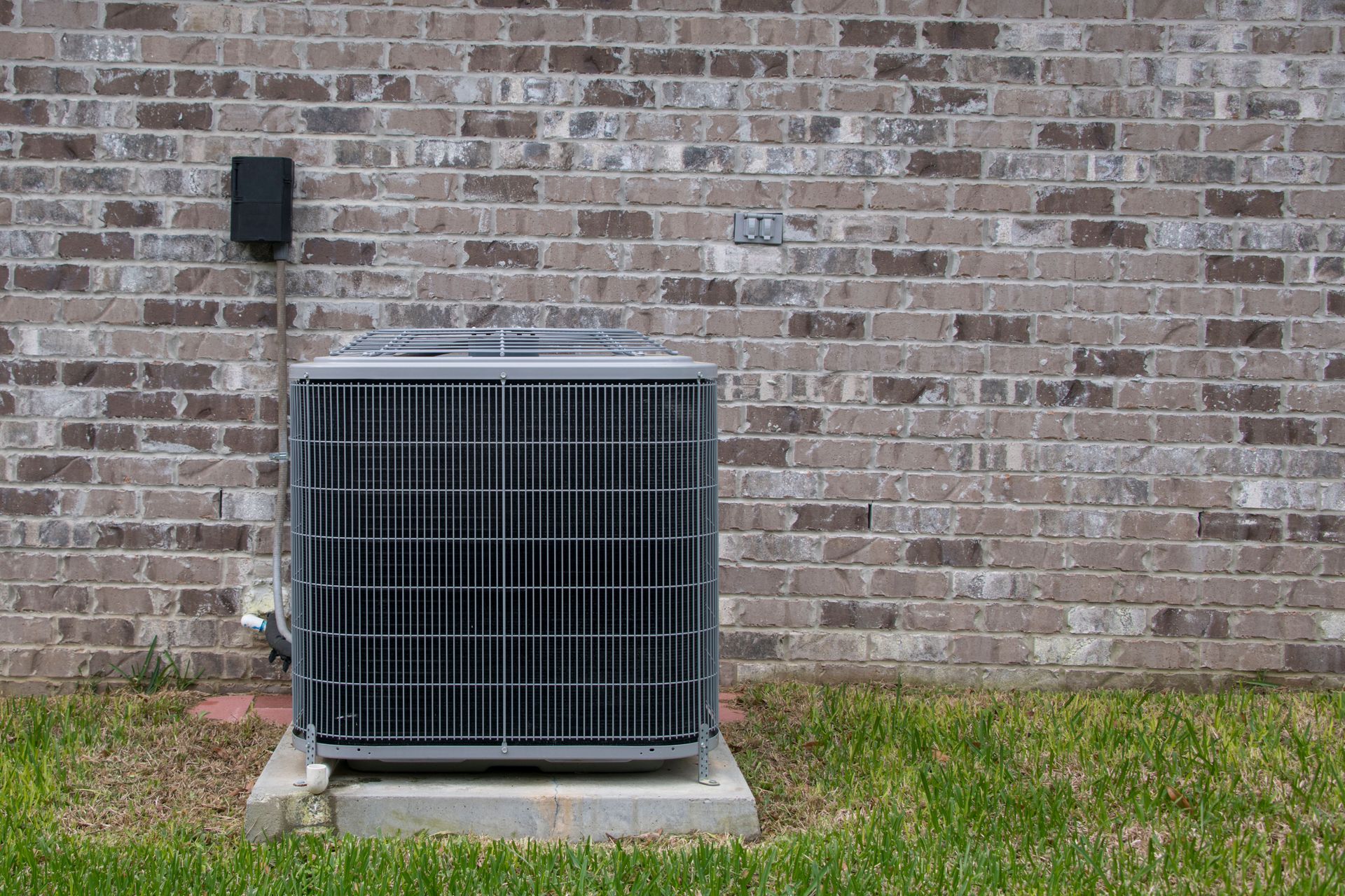 An outdoor air conditioning unit sits on a concrete pad against a brown and gray brick exterior wall with a grassy lawn.