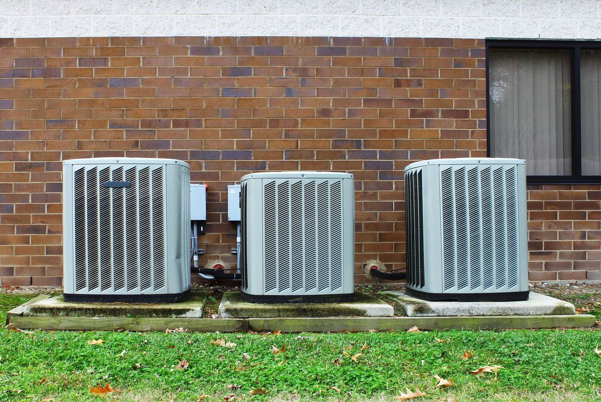 Three light gray outdoor HVAC units sitting on concrete pads against a brown brick building wall.