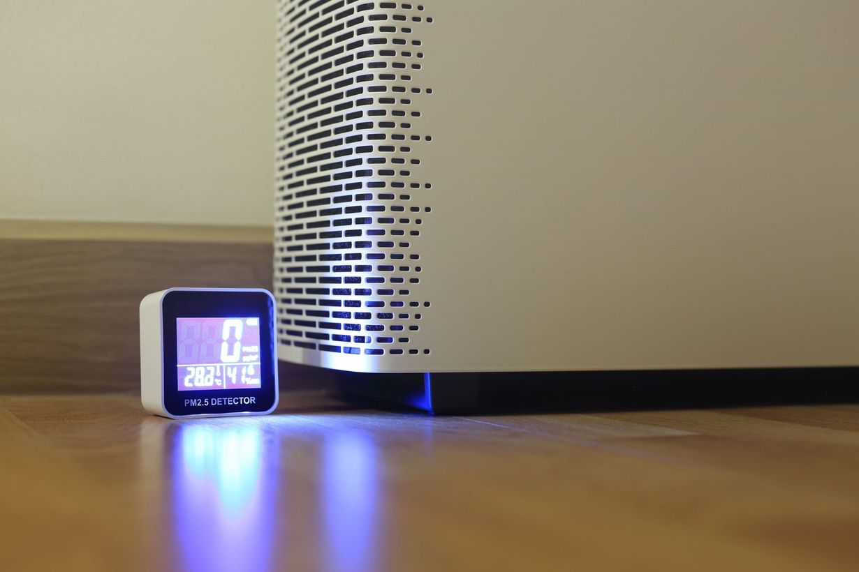 A digital air quality monitor glows blue next to a white air purifier on a hardwood floor.