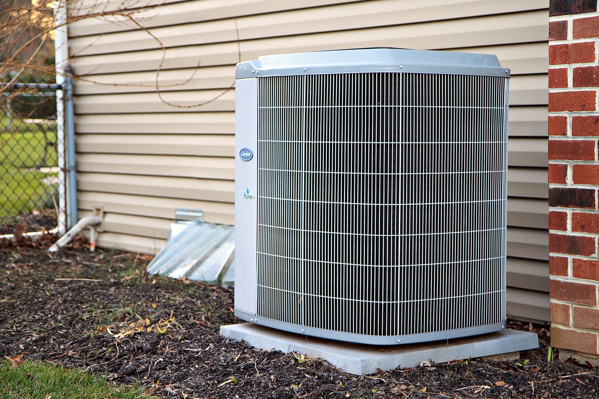 Outdoor air conditioning unit beside a house, with white siding and brick wall backdrop