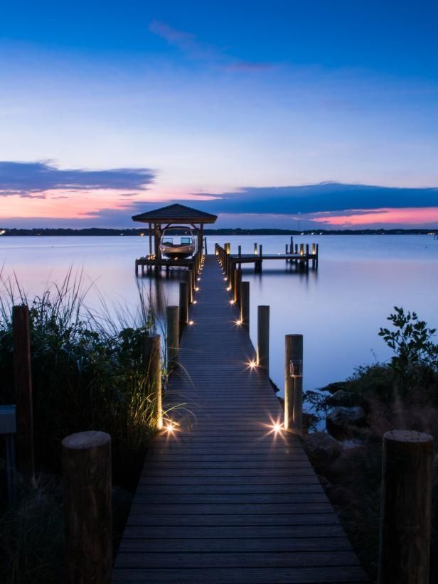 Wooden pier extends over calm water at dusk, illuminated by lights, leading to a gazebo.