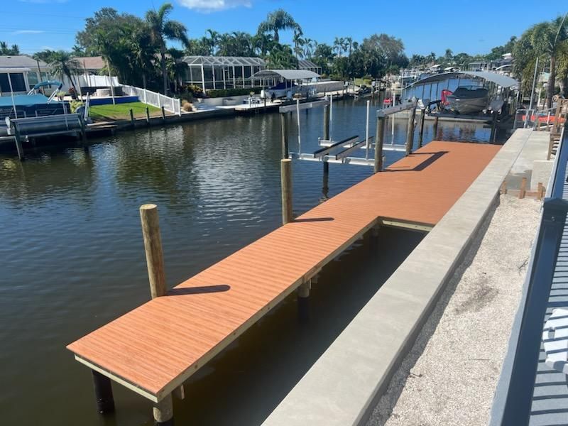 A brown composite dock extends over a canal. Boats and houses are visible in the background on a sunny day.