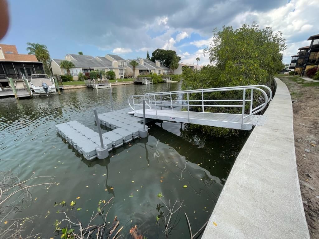 A floating gray dock with handrails on a canal, next to a concrete walkway. Houses in the background.