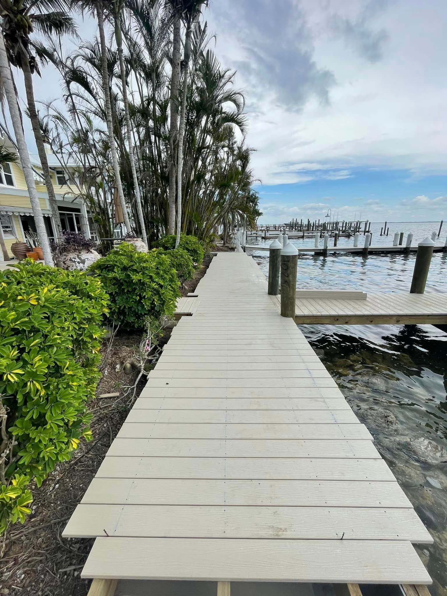 Wooden dock extending to water, lined with shrubs and trees. Cloudy sky.