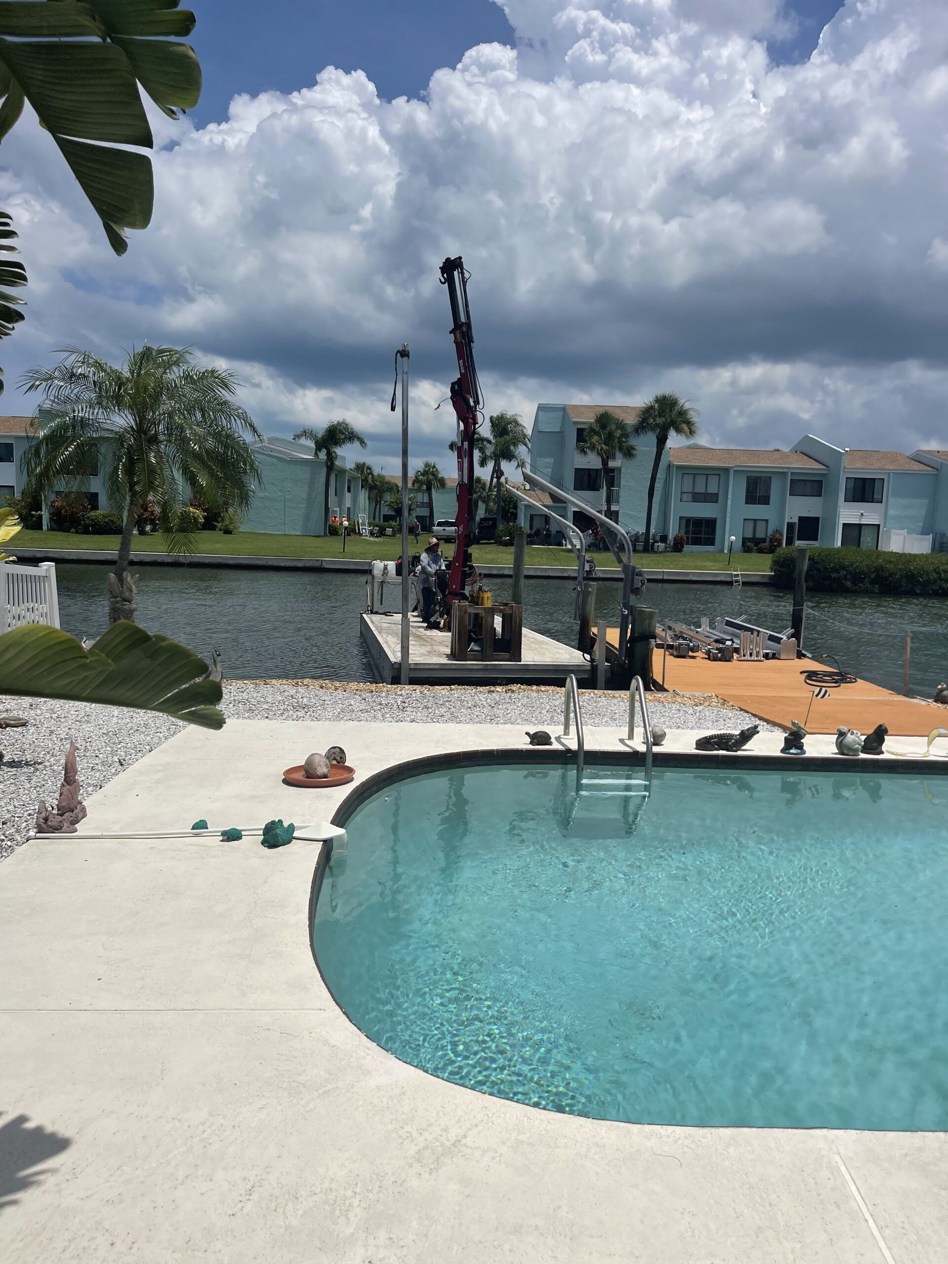 Dock construction beside a pool. A crane lifts materials near turquoise water under a cloudy sky.