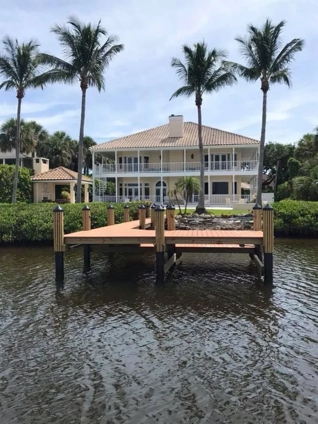 Dock with wooden deck in front of a large beige house with palm trees, set on a canal.