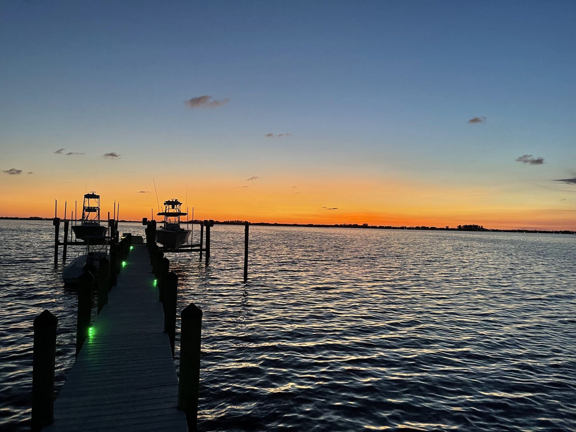 Dock extending into water at sunset, two boats docked, orange and blue sky.