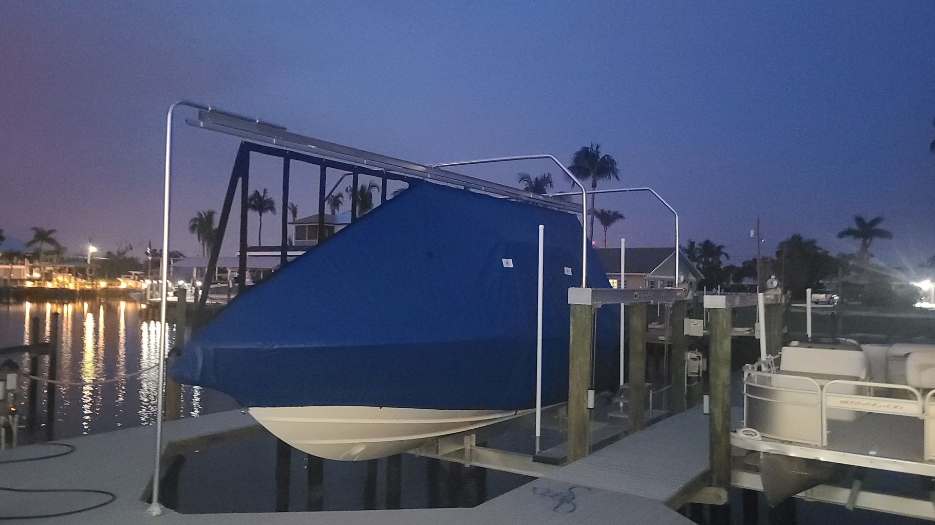 Blue-covered boat on a boat lift at dusk, dock in foreground, lights reflecting on the water.