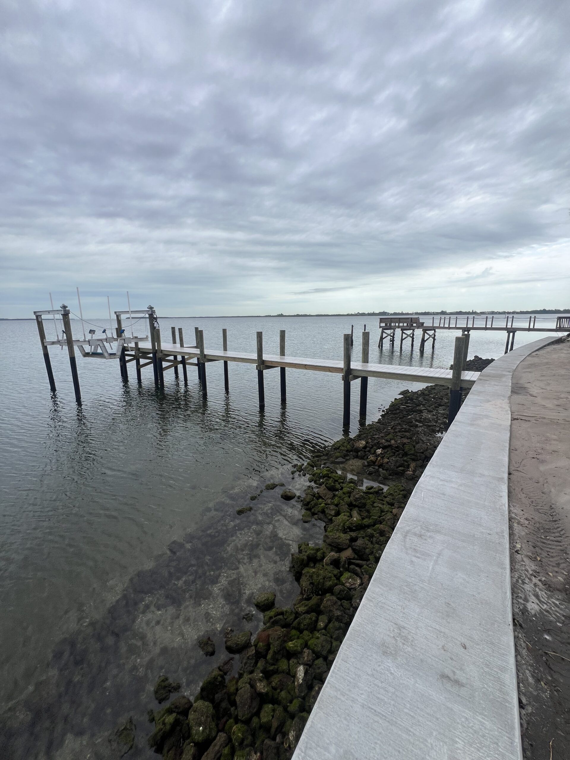 Dock extending into water under a cloudy sky. Shoreline with seaweed and a concrete path on the right.