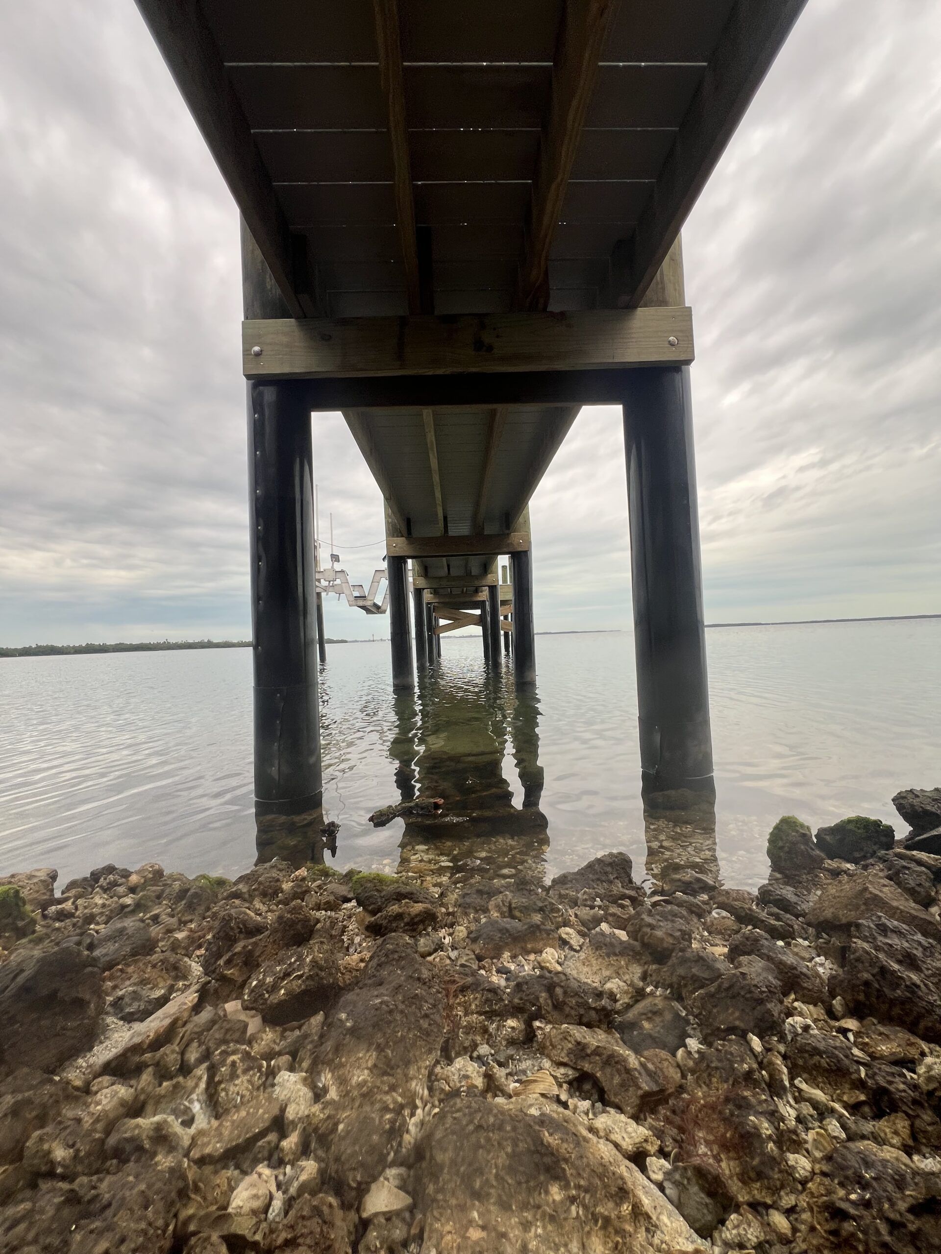 View under a wooden pier, water and rocks in the foreground, cloudy sky overhead.