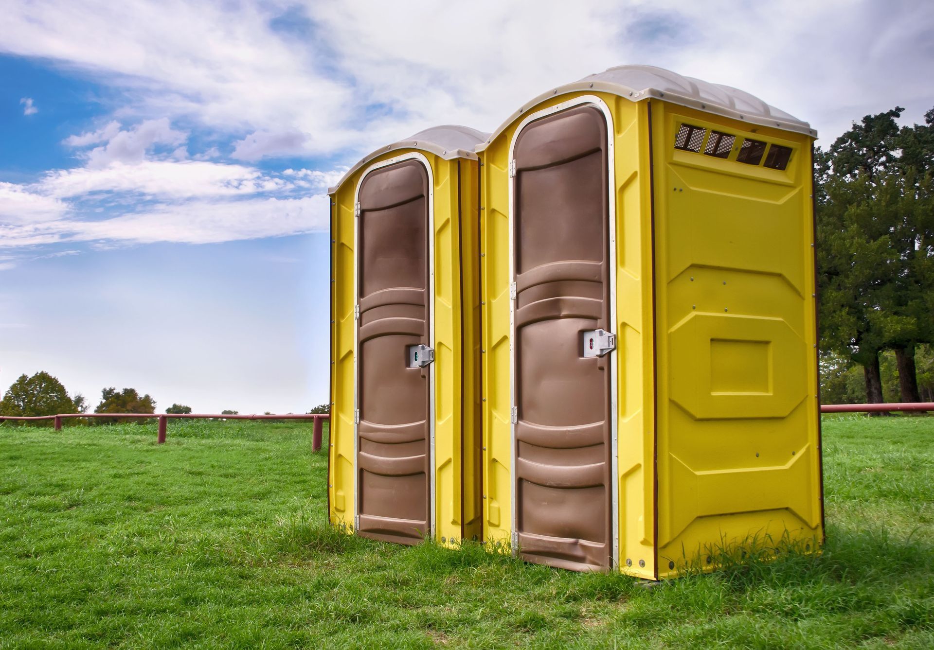 Two yellow portable toilets on green grass, with a blue sky and trees in the background.