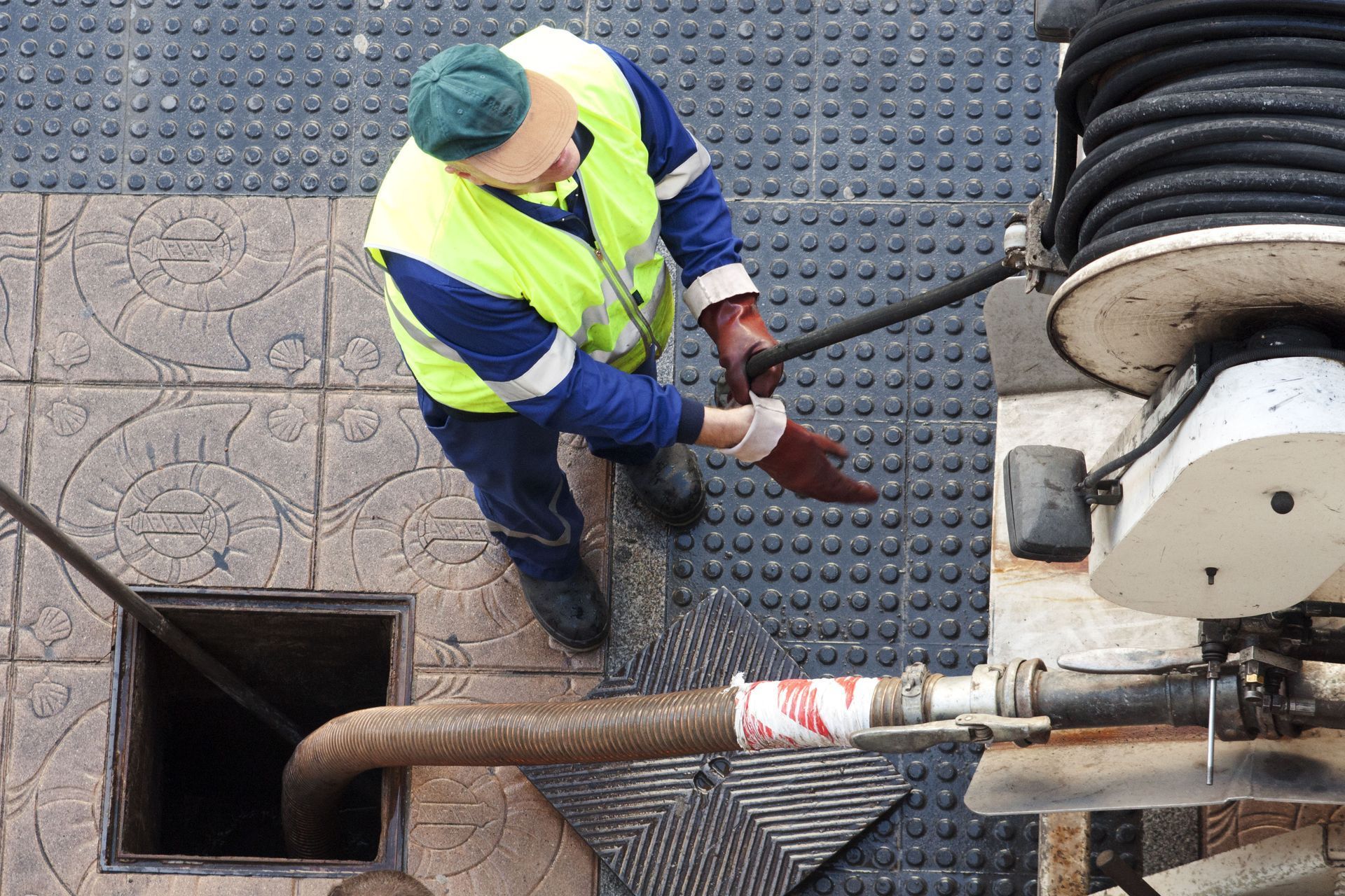 Man in safety vest and gloves working with a hose near an open sewer grate.