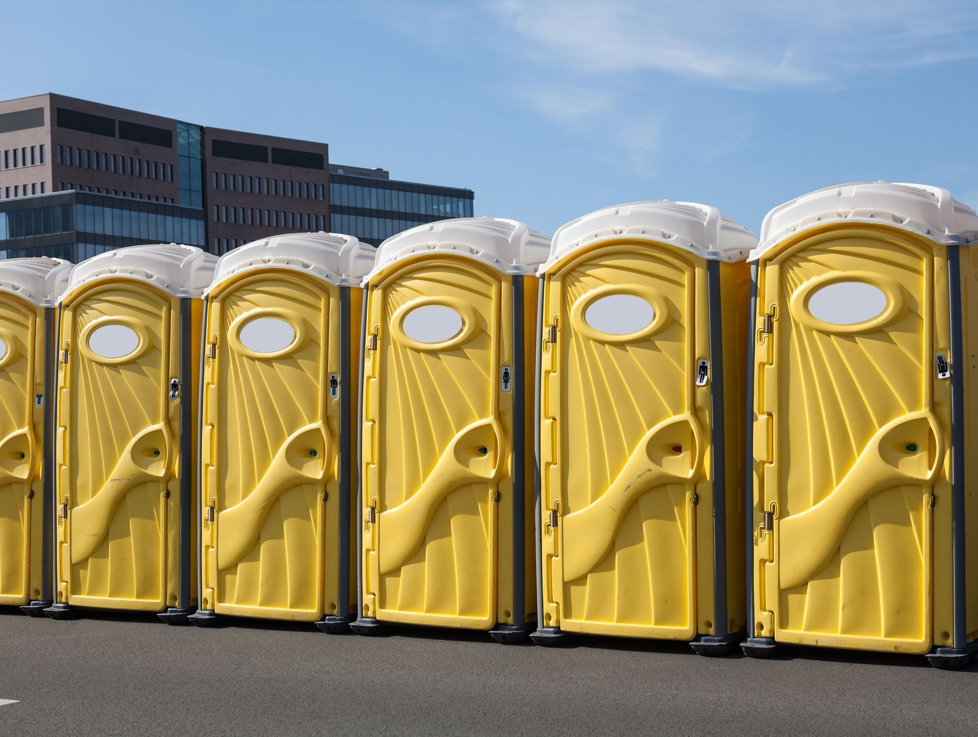 Yellow portable toilets lined up on asphalt, with a modern building in the background on a sunny day.