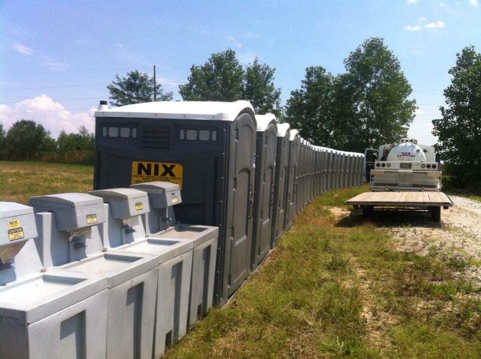 Row of portable toilets and handwashing stations by a grassy area. A truck and trailer are present.