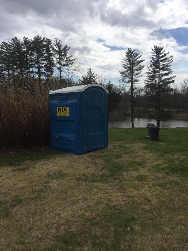 Blue porta-potty on a grassy field, near a pond and tall trees, under a cloudy sky.