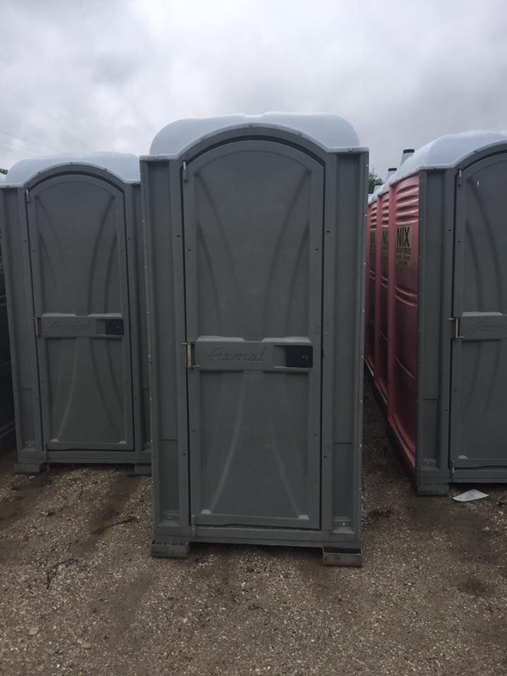 Gray portable toilet, standing outdoors, with two others to the sides; cloudy sky.