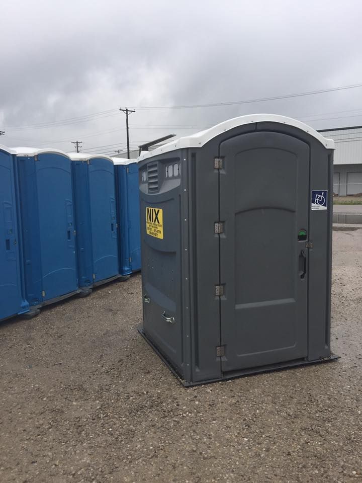 Gray portable toilet in front of a row of blue ones on a wet, overcast day.