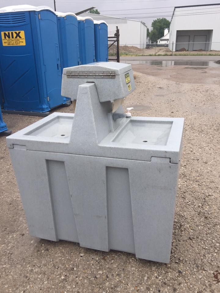 Gray outdoor hand wash station with two sinks, next to blue portable toilets.