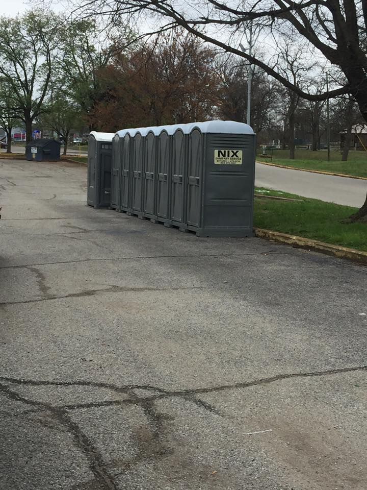 A row of gray portable toilets on asphalt next to a grassy area and trees.