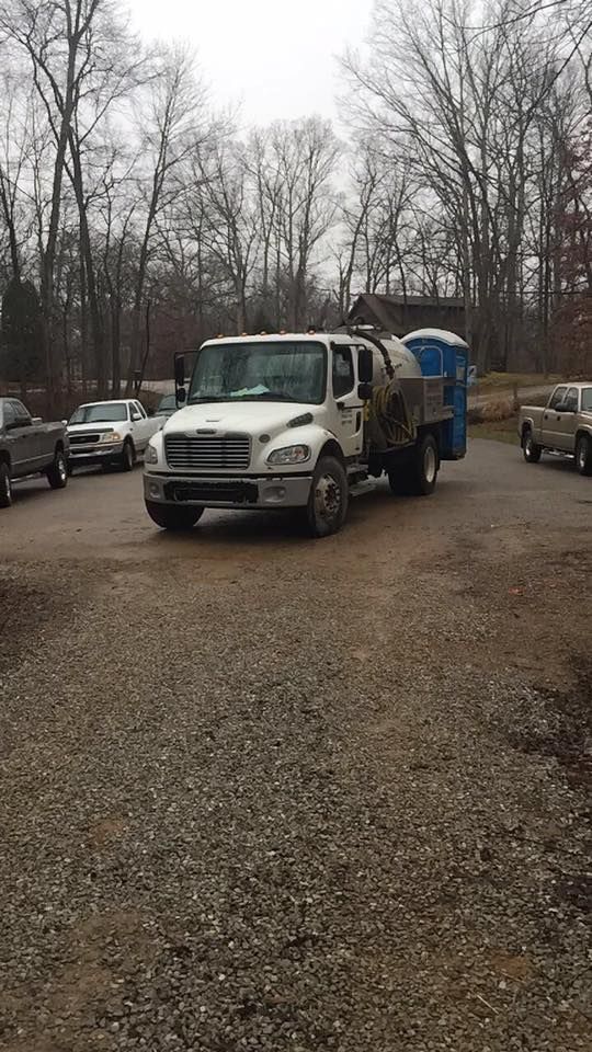 White truck with a portable toilet on the back parked on a gravel lot, trees in the background.