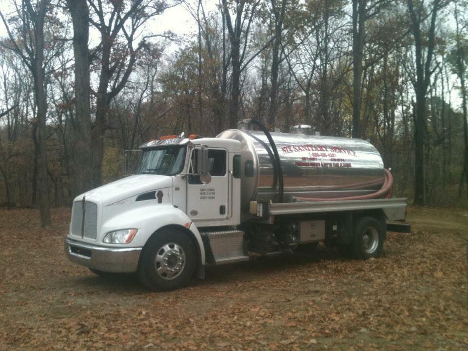 White septic tank truck parked in a wooded area, with a silver tank and company branding.