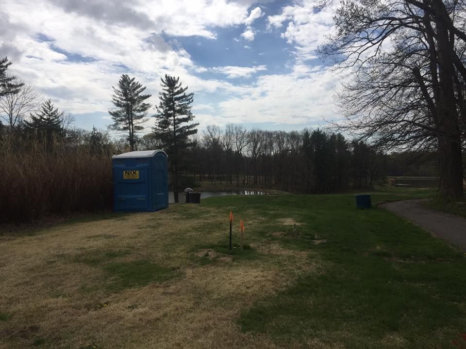 Blue portable toilet on grassy area near path and trees, under cloudy sky.