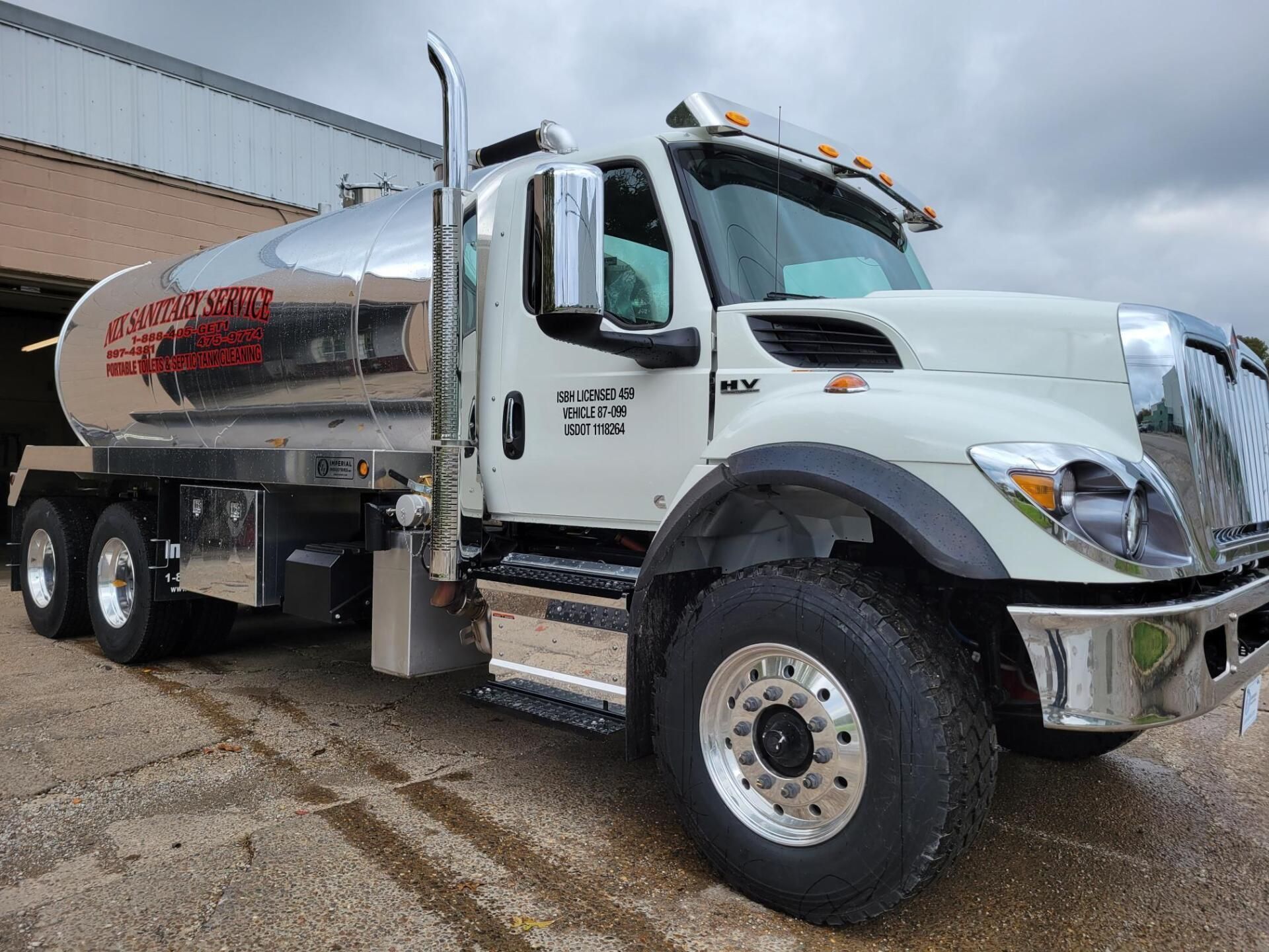 Row of portable toilets and sinks next to a gravel road, with trees and a truck.